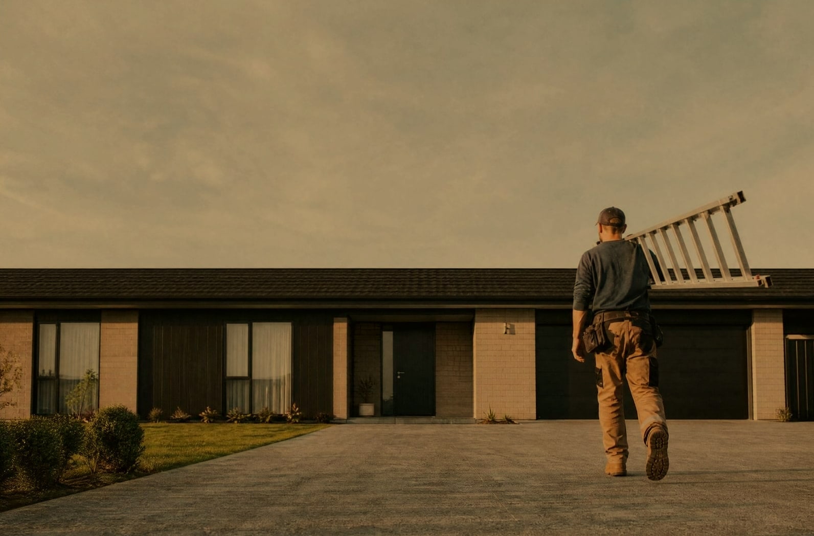 A tradesperson carrying a ladder up a driveway in the early evening, the roofline level across the frame.